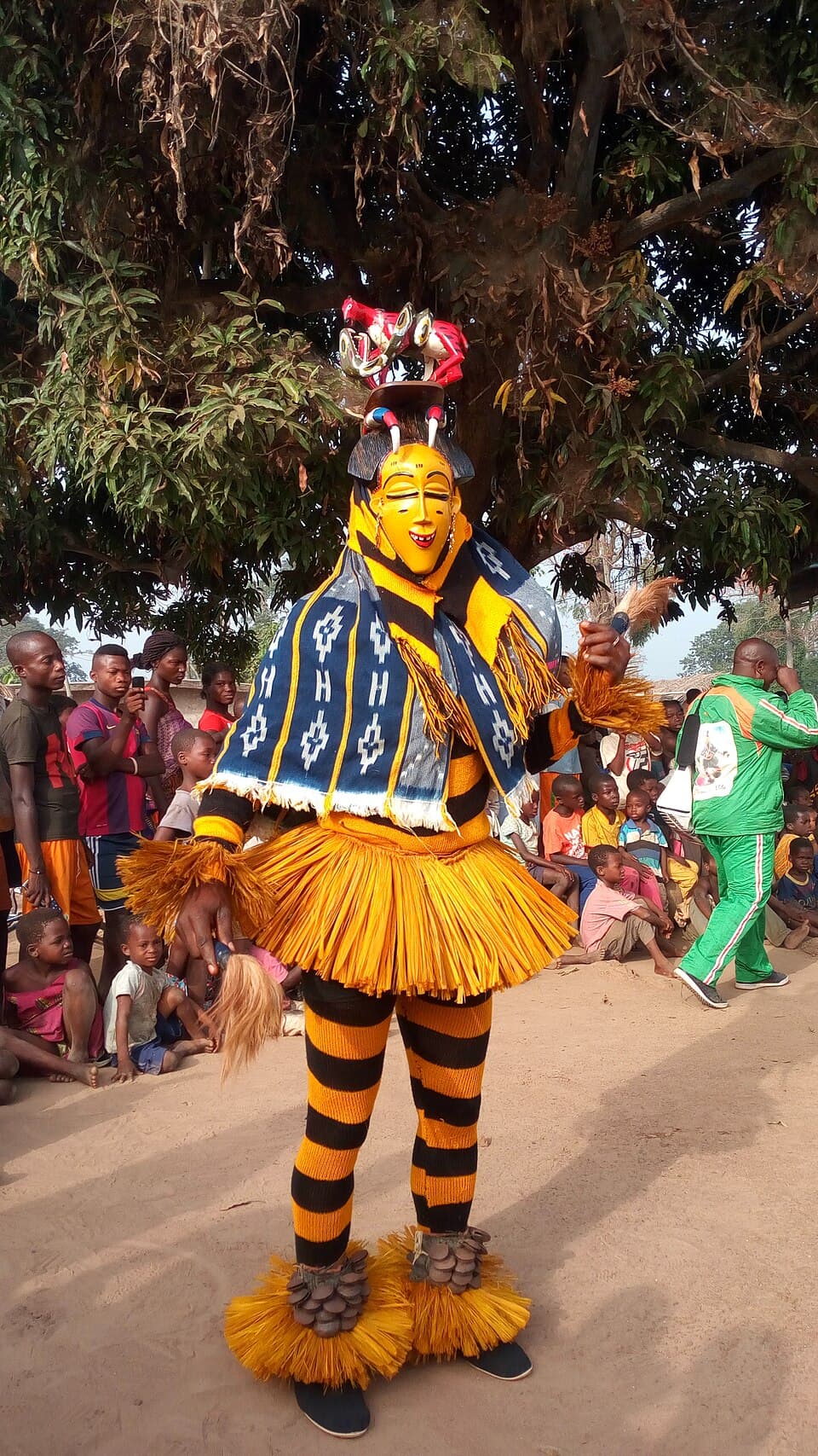 Danseur zaouli en costume complet, masque jaune, coiffe colorée et jupe de raphia, devant une foule à l'ombre d'un arbre